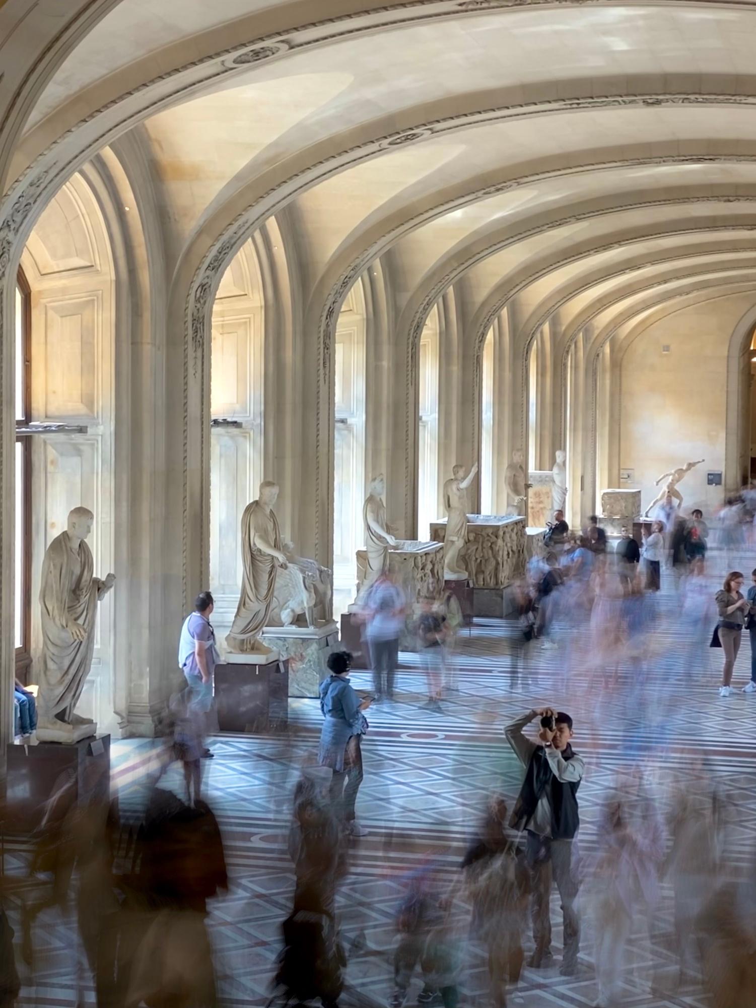 Sala de museu com poucas pessoas, obra em foco desfocado ao fundo.